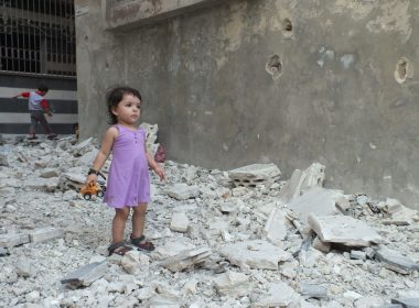 A girl stands on debris next to a damaged building at a besieged area of Homs (HEMA Charity Campaign 2014)